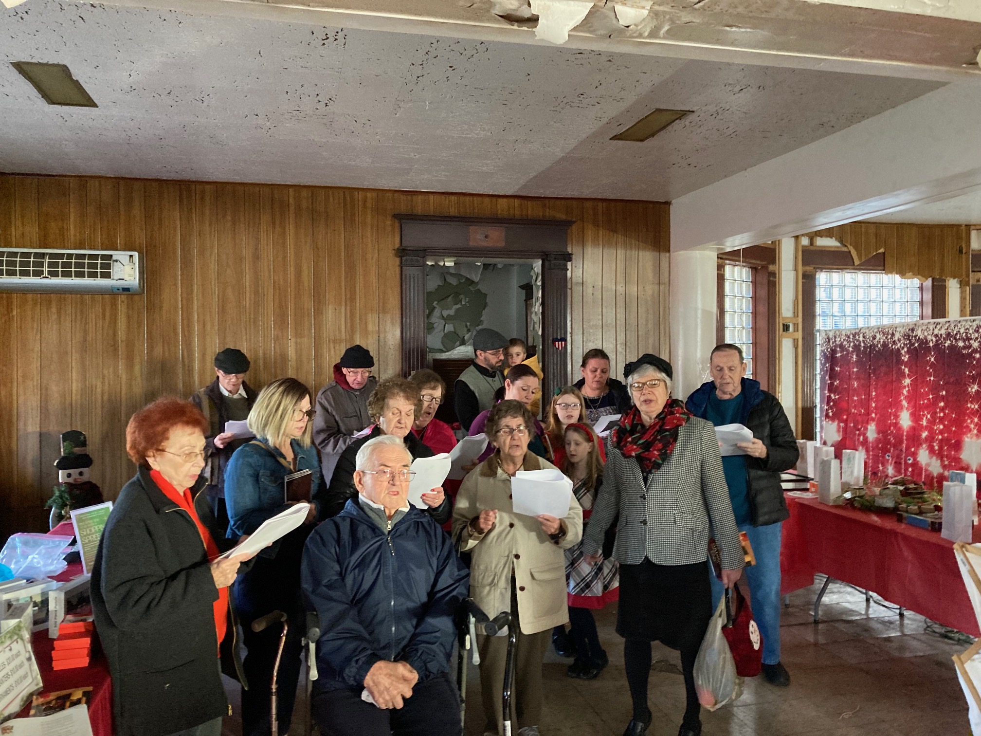 A group of our parishioners singing Christmas carols indoors. There are sixteen people, including three children. The wall behind them is paneled with medium-brown wood, and there's a table on the right side of the frame. The table is covered by a red tablecloth and has white paper bags on top of it. The bags are standing up and are apparently filled with something that is not visible. There are also what appear to be gifts on the table. There's a festive, red screen on the far side of the table that divides the room, apparently to make a cosier space for the event. The screen has big white stars on it that seem to be falling from the top of the screen into rows, leaving a trail of smaller stars in their wake.