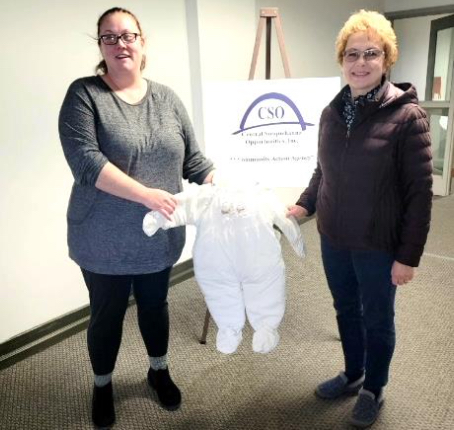 Photo of a CSO representative accepting a white, child-sized, jumper-style coat from Sandy Tosca, president of St. Michael's. The two woman are standing in front of a CSO sign, and each is holding one arm of the coat.