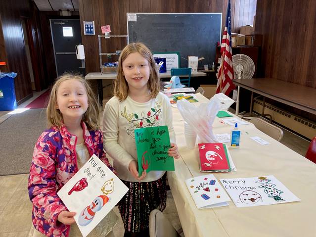 Two girls standing in the church hall, smiling and holding Christmas cards that they created. They appear in the left half of the frame, and there is a long table to their right, running from the right lower corner to the back center of the frame. There are cards and art materials on the table.