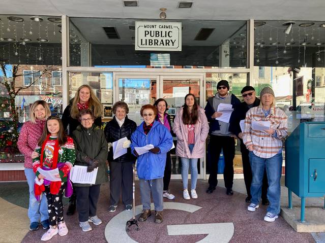 A group of eleven carolers standing in front of a glass storefront, holding sheets of music. A white sign with black letters appears above the glass double-doors and reads 'Mt. Carmel Public Library'. The group consists of a variety of genders and ages, and they're standing on a pink, marble circle with a large 'G' in the center. The library is housed in the former Grossman Department Store, hence the 'G'. Haha, this is bonus information for people with screen readers! :)