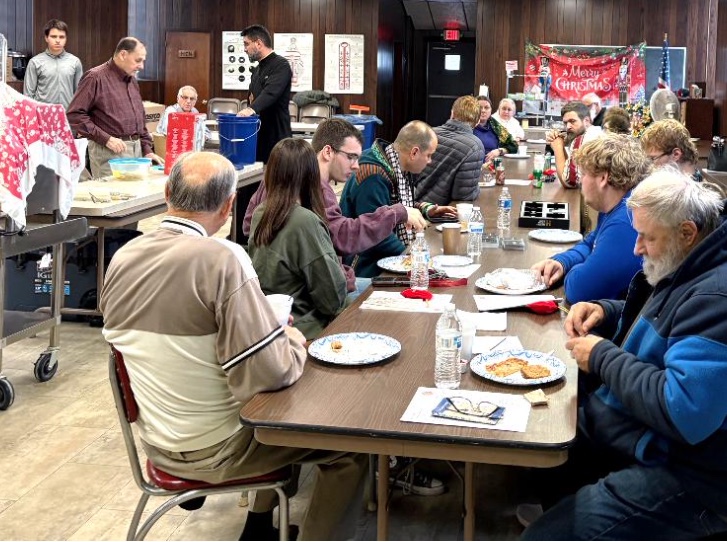 Guests seated at tables and eating in the church hall during the basket raffle. One and a half tables are visible, and Fr. Aleksey can be seen standing with two other people in the upper left corner of the frame as they conduct the raffle.