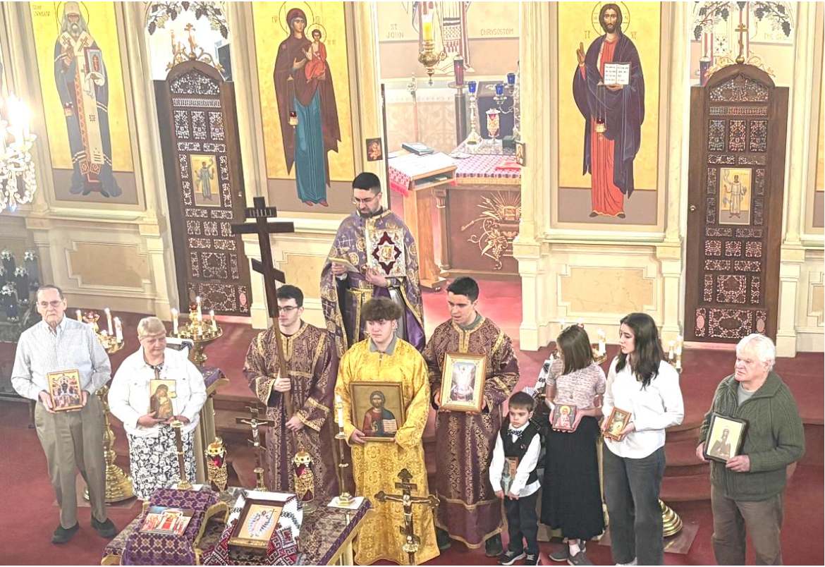 A group of nine people holding icons and posing in front of the iconostasis. One young man is holding a large, wooden cross, and Fr. Aleksey is standing on the steps behind them and is holding a prayer book.