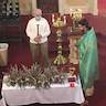 Fr. Vjekoslav, wearing a green vestment, is standing at the upper right corner of a table that is covered with a white table cloth. He is blessing several bunches of pussy willows that are spread across the table. Popadija Dragona is in the lower left of the frame taking a photo. Two men, assisting Fr. Vjekoslav stand on either side of him. The man on the left is holding a candle.