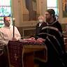 Fr. Vjekoslav is standing in the center of the frame, facing left, towards the (out of frame) altar. His hands are on the bible, which is resting on a lecturn. A male candele-bearer stands to his left, and two parishoners are in pews to the right. A man stands in a front pew, and a woman is kneeling while holding a prayer book.