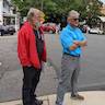 Two men looking to the right at something out of view. The man on the left wears a red jacket and black pants. The man on the right has is arms crossed and is wearing a bright blue shirt, gray pants, and sunglasses.