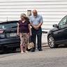 A man and a woman standing behind a car with their backs facing the car. The woman is wearing a plum-colored top and a tan skirt with matching flowers. She is looking to her right. The man is wearing a a light blue shirt and dark gray pants. He has his hands clasped in front of him and is looking down.