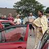 Fr. Vjekoslav is blessing a maroon car that has its driver-side door open while a small crowd of six people looks on.