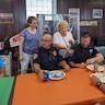 Two police officers are seated at a table with an orange table cloth. They each have a plate of food crumbs and napkins in front of them on the table. There are two women in the photo, standing behind the officers and to their lefts. The woman on the left has a blue, patterned shirt and white pants, and the woman on the right is wearing a white shirt and is reaching for the plate of the officer on the right to take it away.