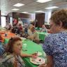 Photo of the church hall during the picnic. Most attendees are seated, and there are two woman talking in the foreground; the one on the left is seated, and the other is standing.