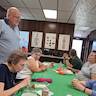 Parishoners seated at a table with a green tablecloth. A man on the left is standing and speaking with a seated man who is on the right.