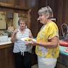 Two women standing in the hall kitchen. The one on the left is wearing a white, sleeveless top with black horizontal stripes and black knee-length shorts. She's pointing or gesturing with her finger pointed. The other woman is holding a plate of food and is wearing a yellow top with white flowers and has on white pants.