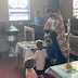 Fr. Vjekoslav, facing to the left of frame, is blessing new, blue vestments that are on a table in front of him. Several rows of pews are shown behind him, and a man, who is holding an open book, is off to Fr. Vjekoslav's right. On Fr. Vjekoslav's left, two young children are observing the blessing of the vestments.