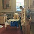 Fr. Vjekoslav, facing to the left of frame, is blessing new, blue vestments that are on a table in front of him. Two pews are shown behind him, and a man is standing off to his right and slightly behind him.