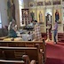 Photo of Fr. Vjekoslav standing in front of a table with fruit on the top. He is holding a prayer book and praying while a male candlebearer stands behind and to his right. A woman on the left side of the frame is taking a photograh, and a young boy stands at the corner of the table, looking off to the right.