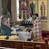 Photo of Fr. Vjekoslav standing in front of a table with fruit on the top. He is holding a prayer book and praying while a male candlebearer stands behind and to his right. A woman on the left side of the frame is taking a photograh, and a young boy stands at the corner of the table, looking at the fruit on the table.