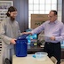 Photo of Fr. Vjekoslav and a parishoner preparing to draw a raffle ticket from a blue, Lowes bucket, which is on an off-white table.
