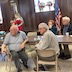 Photo of four parishoners seated at a brown table and looking to the left side of the frame. The two men in the foreground are turned in their chairs, facing away from the table, and a woman and man are on the other side of the table. There's a US flag in a stand behind them. A Popadija Dragona, who is wearing red, is standing in the upper left of the frame, on the far side of the table, and is holding her newborn baby.