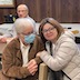 Photo of two women seated in front of a brown table. The woman on the right has shoulder-lenght brown hair, black glasses, an off-white puffer coat, and dark gray pants, and she's looking at the camera. The woman on the left has short, gray hair, and is wearing a brown jacket, light gray pants, black glasses, and a surgical mask. She's looking at the woman on the right. There are two men behind them, on the far side of the table. The man on the left is looking off to the left side of the frame, and the man on the right is looking at the camera.