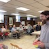 Photo of the church hall during the basket raffle. Fr. Vjekoslav appears on the right side of the frame and is looking at raffle prizes, which appear in the lower left corner of the picture. A group of parishoners are seated at a long brown table in the background and are looking towards Fr. Vjekoslav.