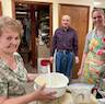 Photo of three people posing for a photo during pascha baking. A woman in the foreground and to the left is holding a mixing bowl with a spatula, and a woman on the right side of the frame stands behind a mixer and with her left hand resting on it. A man wearing a purple shirt and jeans is standing in the center frame and is furthest from the camera.