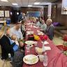 Photo of parishioners seated at a long table in the church hall. The table runs from the front to the back of the photo and is covered in a light red, almost maroon, plastic tablecloth. The people are eating and socializing.