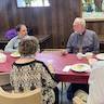 Photo of four parishioners seated at a table covered in a light red, almost maroon, plastic tablecloth. The backs of a woman and man are facing us in the foreground, and two men seated across the table are talking to one another. They are all eating and socializing.