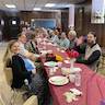 Photo of parishioners seated at a long table in the church hall. The table runs from the front to the back of the photo and is covered in a light red, almost maroon, plastic tablecloth. The people are eating and socializing. Popadija Dragana and Father Vjekoslav are looking towards the camera, and Father Vjekoslav is waving is hand.