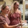 Three women are in the hall kitchen. Two blond women on the left are wearing reddish dresses with flowers. The woman on the left is wearing a clear plastic glove and gesturing with her right hand. A third woman, to the right and further in the background is looking on while wearing a reddish-pink top, tan pants, a light blue surgical mask, and plastic gloves.