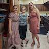 Three blond woman standing in the hall kitchen posing for the camera. The woman in the center is wearing a blue, flowered top and black pants. The other two women are wearing summery dresses. The woman on the left has a pink dress with black dots, and the woman on the right has a flowered pink, orange, and brick red dress.