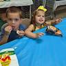 A young boy and girl are sitting at a table eating potatoes chips and looking at the camera. He's at left frame and is wearing a blue shirt while eating plain, Middleswarth chips. She's on the right, wearing a bright yellow dress and bow while eating Lay's barbecue chips. Haha, I love chips! :b