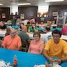 A wideshot of the church hall with guests sitting at tables and talking. Theres are blue tables in the foreground and background and a white table in the middle. Tables run from left to right in the frame, and a woman with great-looking glasses is seated at the table in the foreground, looking into the camera.