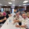 A wideshot of the church hall with guests sitting at tables and talking. Tables run from foreground to background, and the photo mostly shows the people at the center table, which is covered with a white tablecloth. A main at the front of the table is wearing a black-patterned, white shirt and looking into the camera.