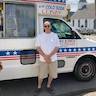 A man in a white t-shirt, tan shorts, and sunglasses is posing with his hands crossed in front of him while standing in front of a white ice cream truck.