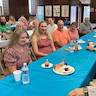 A wideshot of the church hall showing people sitting at long tables and talking. The tables run from foreground to background. The main focus is on a blue-covered table, where in the foreground, a blond woman wearing a pink dress with polka dots is smiling while taring at a cupcake with a candle in it as the others at the table look on.