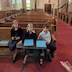 Photo three tweens sitting the front pew during Sunday school. There are two girls on the left, and a boy sitting near the main aisle. The girl in the middle is seated at a small, folding table.