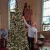 There's a Christmas tree with small, white lights in the center of the photo with a man on the right reaching up to place a ball on the tree. Two cylindrical containers of colored balls are in the foreground, and a man to the left of the tree and towards the back of the photo is opening a small stepladder.