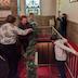 Three woman are decorating the railing that runs around a set of steps heading down to the church hall. The steps are on the right, with the woman on the left. A young girl is standing in the small space between the right-side railing and the right wall, and a young boy is standing at the top of the steps looking towards the landing below. His arms are reaching behind him as he holds onto the railing.