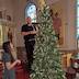 A man dressed all in black is standing on a short stepladder and wrapping ribbon around a Christmas tree. A woman in a black dress with white polka dots is standing in the lower, right corner of the photo and is watching.