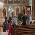 Six people are decorating a Christmas tree and/or watching. There are several rows of pews in the foreground.