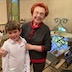 A boy in a white shirt is posing with a woman wearing dark gray with red trim. Both are smiling, and a table and some chairs appear in the background with books on them.