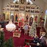 A view of the church from the choir loft. The priest is standing behind a lecturn draped in a red cloth with a cross, and he's facing the congregation. The first few pews on each side are visible, with some people standing within them. A Christmas tree is located in front of the pews on the right, and a man holding candles is standing to the left of the tree and is facing the iconostas. Evergreen garland that runs along the choir loft railing can be seen in the lower left corner.