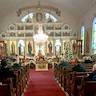 A view from the back of the church, looking down the aisle, between the pews. The iconostas is shown, and the priest can be seen through the Royal Doors. There's a Christmas tree in front of the pews on the right.