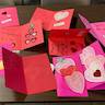 Photo of handemade Valentine's Day cards on a brown table. The tops of two folding chairs are visible on the far side of the table. Cards are red and pink, with hearts. A couple have lace hearts. Some cards are open, with inside wishes such as 'Happy Valentine's Day and 'You're the best'.