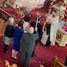 The wedding couple stands before Father Vjekoslav, who is holding a book. Two women and a man are standing behind the couple, and the women on the left and the man on the right are holding crown above the heads of the wedding couple. The crowns have rods attached, and the crown bearers are holding the rods such that the crowns are hovering above the couple's heads.