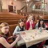 Five students (four girls and a boy) are seated around a folding table set up in front of the first pew in the church. Their female instructor is crouched down in front of the table and looks on as they work on decorating their crosses.