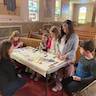 Five students (four girls and a boy) are seated around a folding table set up in front of the first pew in the church. Their female instructor is crouched down in front of the table and looks on as they work on decorating their crosses.