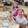 Photo of the pussy willow project at Sunday school in the church hall. The teacher, a woman wearing a black, long-sleeved top, is seated on the left side of a long table with two young girls seated across from her. The girls are looking at the camera and smiling. There are pussy willows, paper plates, picture frames, and other craft materials on the table, which has an off-white table cover on it.