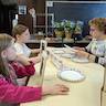 Photo from Sunday school class, where two young girls are seated on the left-hand side of a long table that is covered with an off-white table cloth. They're both holding off-white picture frames. They are listening to a teacher, who is seated across the table from them and is reading to the girls. There are paper plates and small bottles of paint on the table.