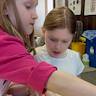 A close-up shot of two young girls working on their craft projects. The girl in front is reaching across the bottom front of the frame for something off-camera.