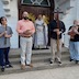 Photo Fr. Barnabas standing at the outside front of the church, flanked by two males on either side, each holding one item. From left to right, the males are holding a candle, a bowl, a large wooden cross, and a Holy Bible, while the priest holds a gold cross.